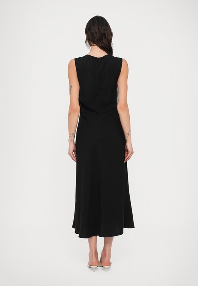 Sieviete melnā, bezroku, garā kleitā stāv ar muguru pret kameru vienkāršā baltā fona priekšā.