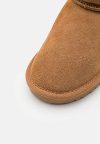 Bottine en daim marron avec un bout arrondi, une surface texturée et une semelle en caoutchouc flexible. Détails de couture près du bord supérieur.