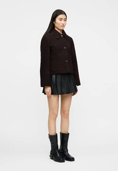 Γυναίκα όρθια, που φοράει σκούρο καφέ σακάκι με κουμπιά, μαύρη πλισέ μίνι φούστα και μαύρες δερμάτινες μπότες μέχρι τη μέση της γάμπας, σε λευκό φόντο.