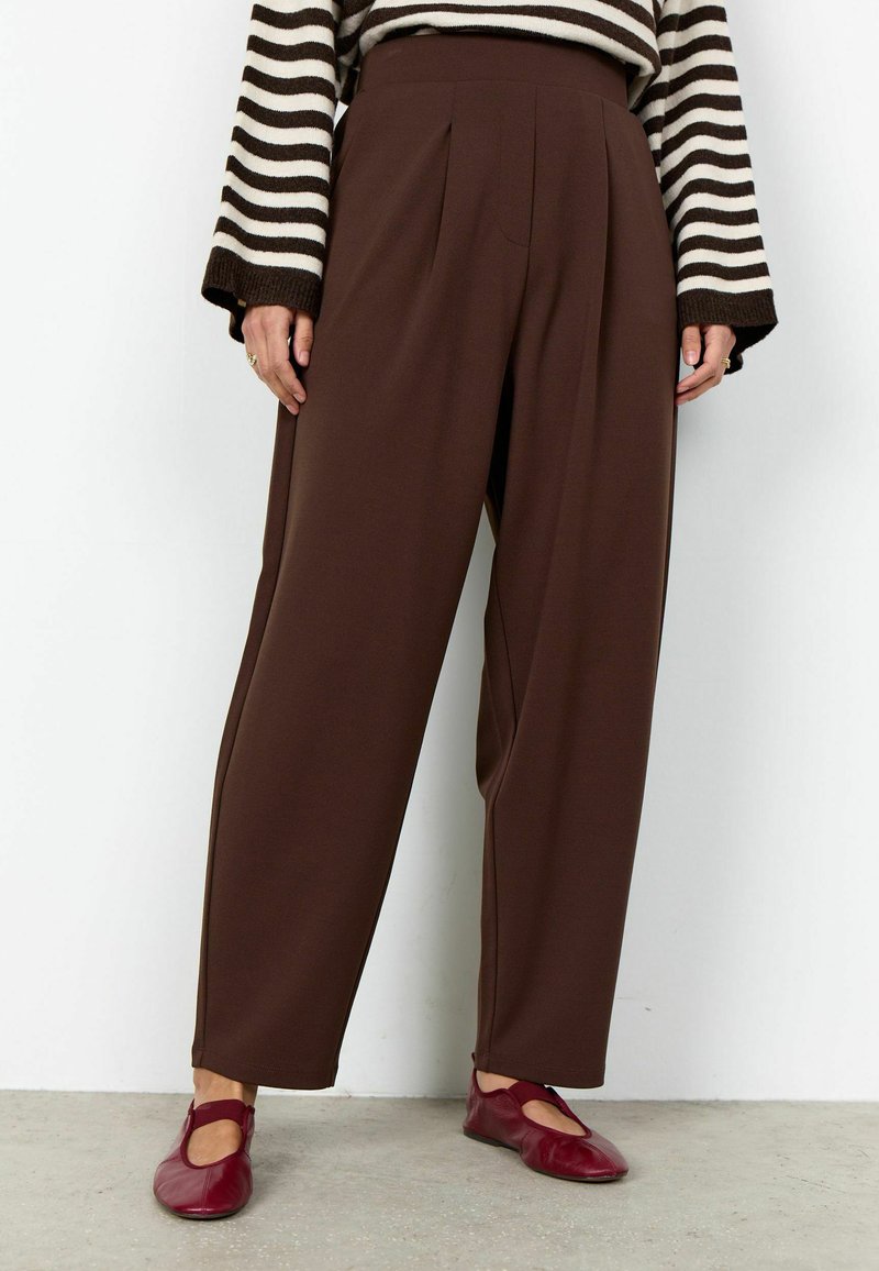 Persoană purtând pantaloni maro cu talie înaltă și croială largă, bluză cu mâneci lungi cu dungi, și pantofi roșii fără toc, stând pe un podea gri.