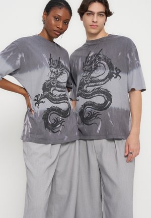 Zwei Personen tragen passende graue Batik-T-Shirts mit großen schwarzen Drachendesigns, kombiniert mit locker sitzenden hellgrauen Hosen.