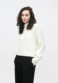 Jersey de cuello alto de punto grueso en color crema, con puños y dobladillo acanalados, combinado con pantalones negros de tiro alto, mostrando un ajuste holgado.