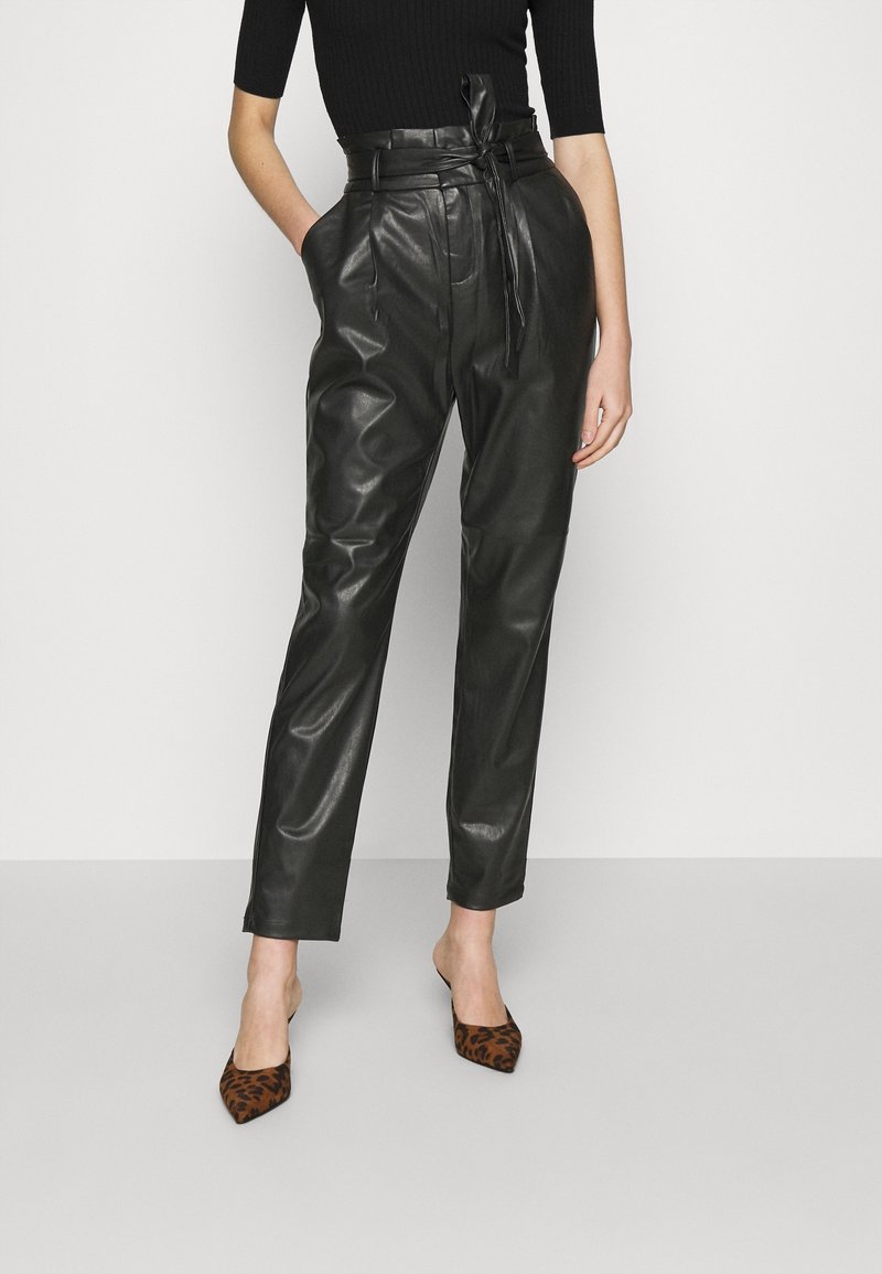 Femme portant un pantalon en cuir noir taille haute avec ceinture nouée, un haut noir et des talons pointus imprimé léopard, debout les mains dans les poches.