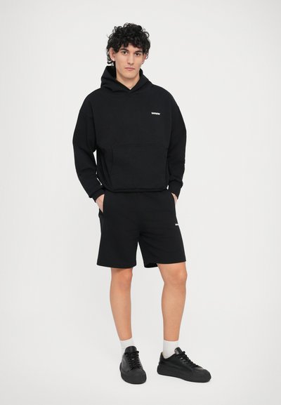 Sweat à capuche en coton noir avec poche avant, associé à un short noir. Des baskets noires et des chaussettes visibles à la cheville complètent la tenue.