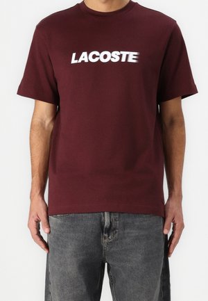 Mężczyzna ubrany w ciemnoczerwoną koszulkę z białym napisem "LACOSTE" i wyblakłymi czarnymi dżinsami, stojący na jednolitym jasnym tle.