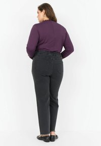 Kobieta odwrócona tyłem, ubrana w fioletową bluzkę z długim rękawem, czarne jeansy z wysokim stanem i elastycznym pasem oraz czarne mokasyny na białym tle.