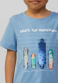 Kind in einem blauen Shirt mit Pailletten und bedruckten Skateboards, die mit "Slide", "Slalom", "Speedboard" und "Classic" beschriftet sind, vor einem Höhenmessstab-Hintergrund.