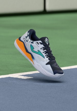 Scarpa sportiva con accenti bianchi, blu navy, verde acqua e arancioni sospesa sopra un campo da tennis vicino alla linea di fondo su una superficie verde e blu.