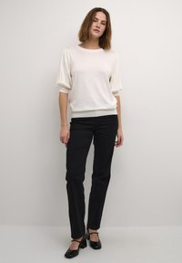 Pull en laine léger de couleur crème avec des manches courtes bouffantes, associé à un jean noir slim et des chaussures à talons carrés noires. Design minimaliste.