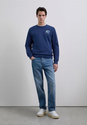 Marineblauwe sweatshirt met logo op de borst, gecombineerd met lichtblauwe jeans. Model draagt witte sneakers met gele zolen. Eenvoudig, casual ontwerp.