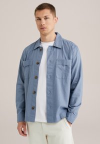 Camisa de botones azul claro con cuello extendido, dos bolsillos frontales y botones marrones. Hecha de tela texturizada, se lleva sobre una camiseta blanca.