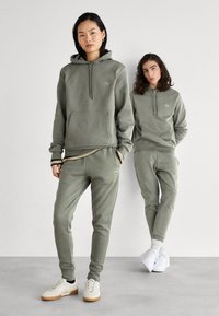 Sudadera de algodón verde con logotipo, bolsillo frontal, cordones; combinada con pantalones de chándal a juego. Tejido texturizado, corte relajado, zapatillas blancas.