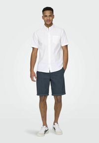 Chemise blanche à manches courtes avec une poche poitrine, assortie à un short gris foncé et des baskets blanches avec des accents noirs.