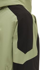 Jakke med tofarvet design, hvor kroppen er lys grøn og skuldrene har sorte accenter, lavet af et glat, holdbart stof.