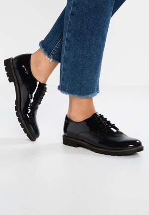 Sorte skinnende snøresko båret med blå jeans med frynset kant, den ene fod fladt på gulvet, den anden let løftet på tæerne.