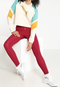 Felpa corta con pannelli multicolori in crema, teal e arancione; abbinata a leggings rossi e sneakers bianche con dettagli in rete.