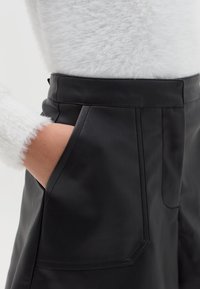 Pantaloni corti in pelle nera a vita alta, con tasche laterali e dettagli di cucitura visibili. Manica di un morbido maglione bianco parzialmente visibile.