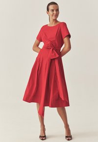 Piros pamutruha rövid ujjakkal, fitted derékkal, masnival megkötve, bő szoknyával, redőkkel ellátva és egyszínű. Fekete nyitott orrú cipővel viselve.