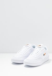 Paire de baskets basses blanches Nike avec un bout perforé, une fermeture à lacets à l'avant, et le logo Nike orange-noir sur la languette et le côté, sur une surface blanche.