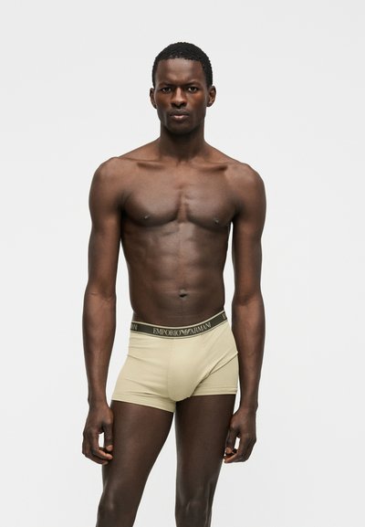 Αθλητικός άνδρας με σκουρόχρωμη επιδερμίδα, ο οποίος στέκεται και κοιτάζει μπροστά φορώντας μπεζ boxer briefs Emporio Armani, πάνω σε λευκό φόντο.