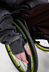 Pelēks džemperis ar ribotām aprocēm, daļēji aptver roku, kas tur melnu ratiņu ratu ar spilgti zaļu apmalu.