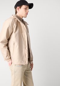 Beige lättviktsjacka med huva, dragkedjefickor, elastiska muddar och en mjuk textur. Tillsammans med ljusa byxor.