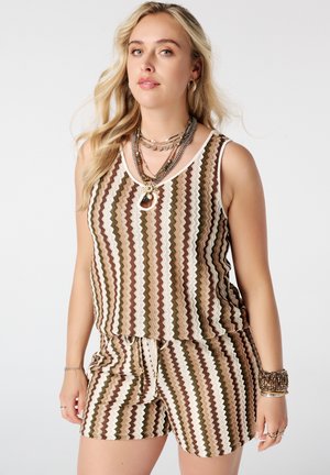 Vrouw met een mouwloze top en short met golvende bruine, crèmekleurige en olijfgroene strepen, laagjes kettingen en armbanden, staand tegen een effen achtergrond.