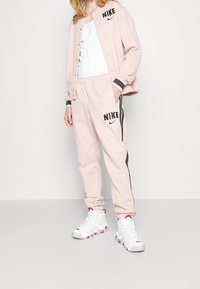 Rosa Nike träningsoverall med svarta accenter; jacka med tryckknappar, byxor med elastisk midja, med logodetaljer. Vita sneakers fulländar outfiten.