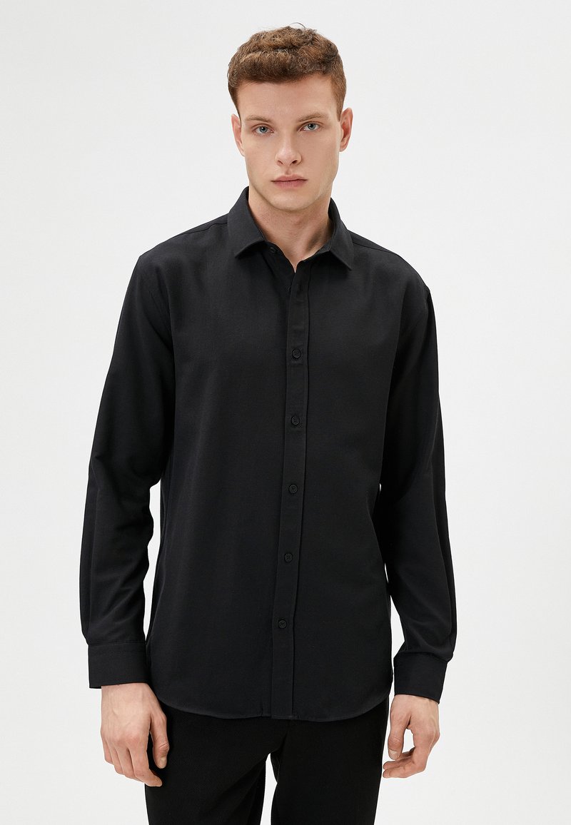 Camisa negra de manga larga con cierre de botones en la parte delantera, cuello clásico y textura suave. Presenta un corte ajustado y puños con un solo botón.