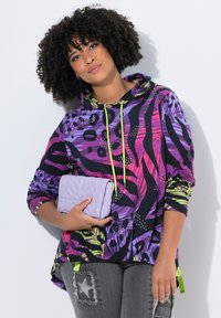 Mujer con cabello rizado que lleva una sudadera con un patrón vibrante en púrpura, rosa y verde, y sostiene un bolso de mano acolchado en color lavanda.