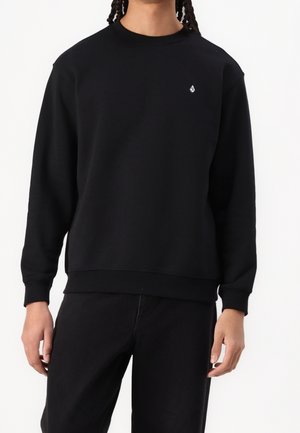Personne portant un sweat-shirt noir uni à manches longues avec un petit logo blanc sur la partie supérieure de la poitrine et un pantalon noir, sur fond blanc.