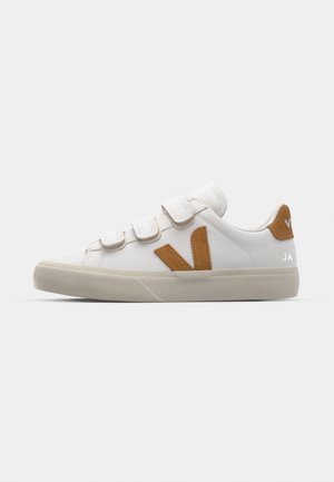 Hvide sneakers med brune ruskindsdetaljer, tre velcro-stropper og en tekstureret gummisål. Har logo på hælen. Simpelt, atletisk design.