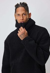 Pull-over en polaire noire à col haut avec une demi-fermeture éclair, manches longues et poches avant. Extérieur texturé et coupe décontractée.