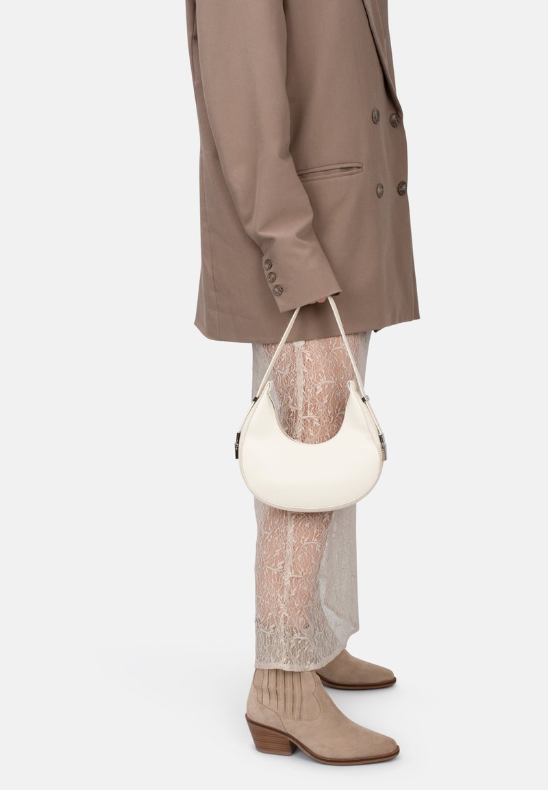 Borsa in pelle bianca con forma curva, tenuta con un manico corto; abbinata a un blazer beige e pantaloni in pizzo, completata da stivaletti alla caviglia color marrone.
