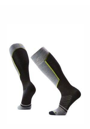 Par de calcetines deportivos negros y grises hasta la rodilla con detalles amarillos exhibidos en piernas de maniquí en pose de caminar.