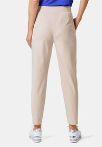 Beige Slim-Fit-Hosen mit glatter Textur, ausgestattet mit einem nahtlosen Bund und zwei Gesäßtaschen, kombiniert mit weißen und silbernen Sneakern.