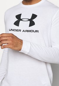 Biała koszula z długim rękawem, wykonana z miękkiego materiału, z dużym czarnym logo Under Armour na piersi. Prosty krój z okrągłym dekoltem.