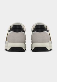 Ζευγάρι λευκών και γκρι αθλητικών παπουτσιών με μαύρες ταμπέλες στη φτέρνα και χρυσές καρδιές ως διακοσμητικά, παρουσιασμένα από πίσω σε απλό φόντο.