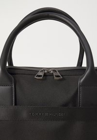 Schwarze Nylon-Handtasche mit Lederakzenten, ausgestattet mit zwei runden Griffen, einem Reißverschlussverschluss und einem Logo auf der Vorderseite.