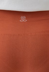 Pružné, žebrované legíny v barvě rzi s hladkým povrchem, s bílým logem na pase jako jemný branding detail.