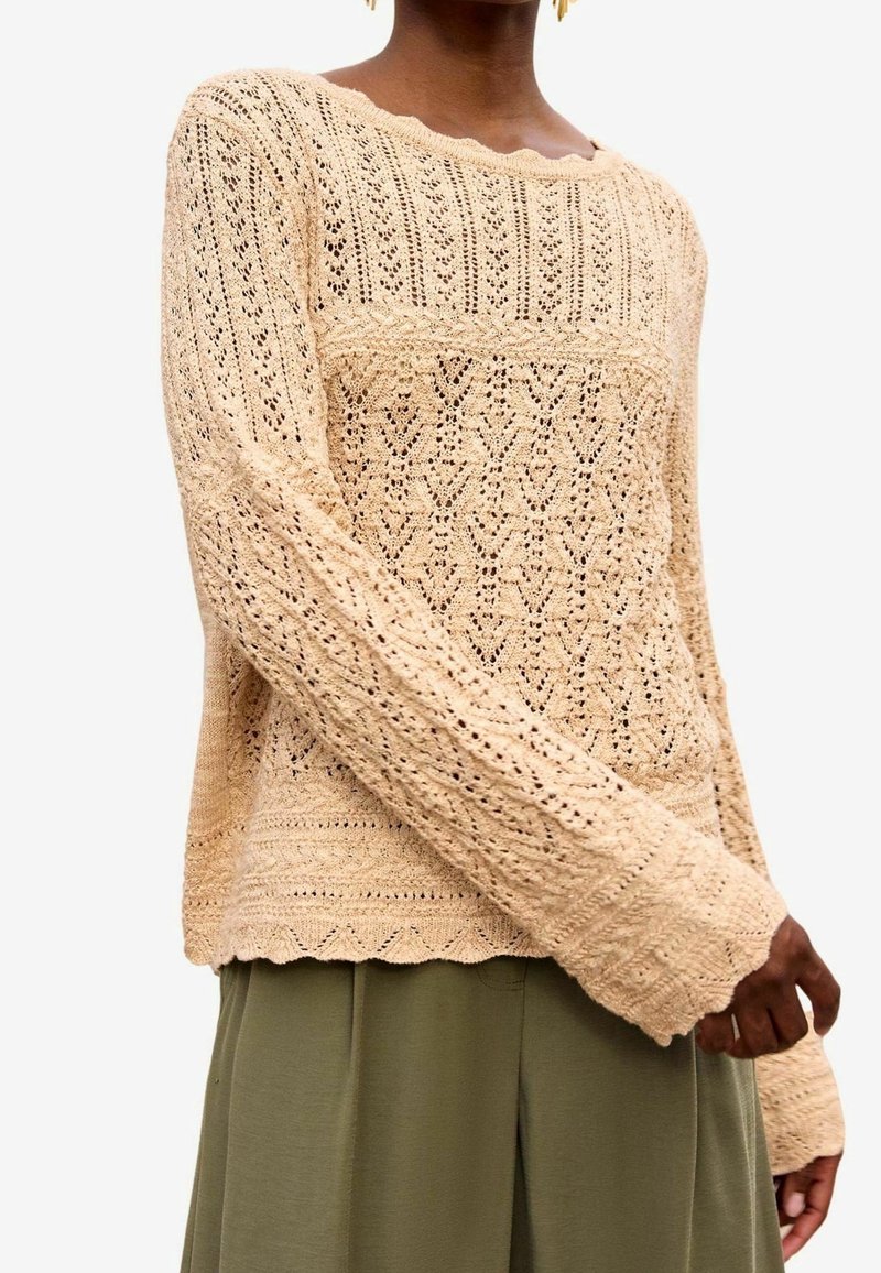 Pull en tricot de couleur beige clair, doté de motifs ajourés, de manches longues, d'un ourlet festonné et de détails texturés tout au long.