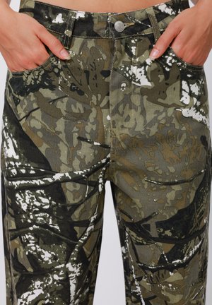 Hænder stukket i lommerne på camouflage-mønstrede bukser med knap og lynlås foran.