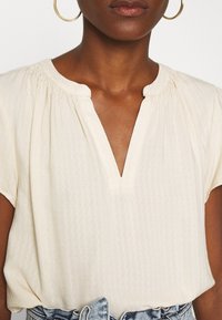 Krémová blůza s krátkým rukávem, výstřihem do V a texturovaným vzorem ve tvaru cik-cak, s nařasenými detaily kolem krku. Spárovaná s džínovými šortkami.