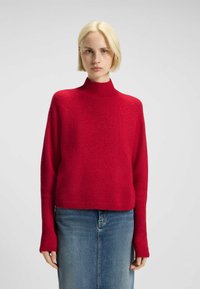 Pull rouge à col haut avec une coupe décontractée, des manches longues et des poignets côtelés, fabriqué à partir d'un tissu doux et texturé. Porté sur une jupe en denim.
