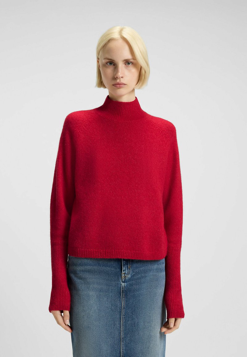 Pull rouge à col haut avec une coupe décontractée, des manches longues et des poignets côtelés, fabriqué à partir d'un tissu doux et texturé. Porté sur une jupe en denim.
