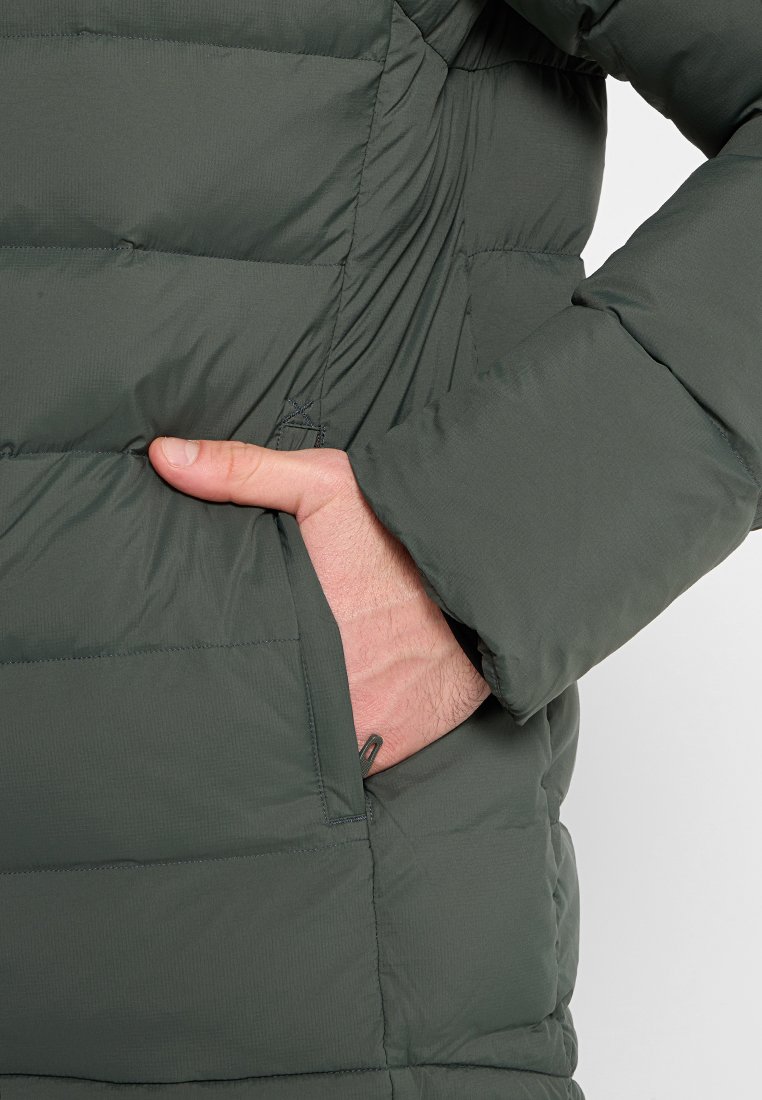 Una mano inserita nella tasca laterale di un giubbotto imbottito verde scuro con dettagli di cucitura orizzontale.