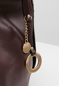 Gros plan d’un sac en cuir brun foncé texturé avec des breloques dorées circulaires et en forme de disque gravées "ANNA FIELD."