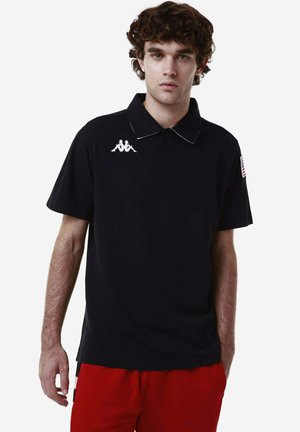 Polo shirt nera a maniche corte, con bordatura del colletto bianca e logo Kappa sul petto sinistro; abbinata a pantaloni corti rossi. Tessuto di cotone liscio.