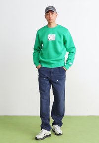 Felpa verde con stampa grafica, jeans blu e sneakers bianche. Modello in piedi con le mani in tasca su uno sfondo semplice.