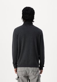 Uomo con capelli scuri che indossa un maglione grigio scuro a collo alto e pantaloni cargo grigi chiari, visto di spalle su uno sfondo bianco.